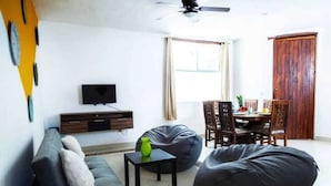 Ein Wohnzimmer mit einer Couch, zwei Sitzsäcken, einem kleinen Tisch mit einer Vase, einem Fernseher auf einem Holzgestell und einem Essbereich mit Stühlen und einem Tisch.