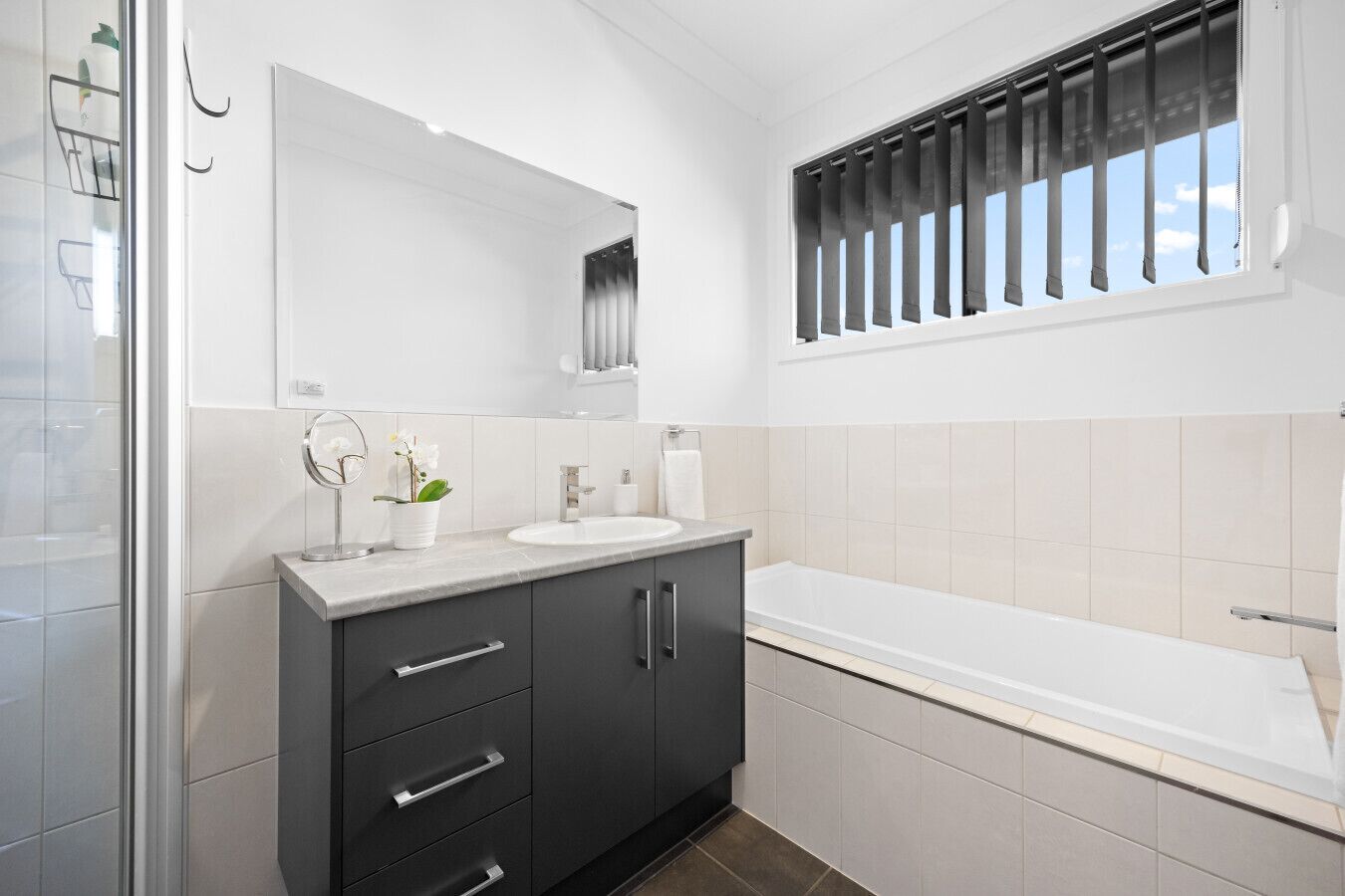 A modern bathroom with a large mirror, a sink, a vanity with drawers, a bathtub, and a window with blinds.