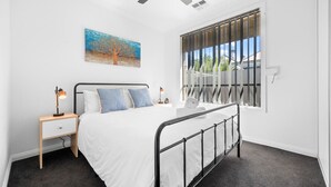 Un dormitorio con cama, mesita de noche, lámpara de escritorio, ventilador de techo, ventana con persianas y un cuadro en la pared.