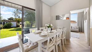 Uma sala de jantar com mesa posta para seis pessoas, cadeiras brancas e vista para o jardim através de amplas janelas.