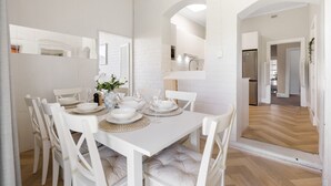 Uma área de refeições com uma mesa branca para seis pessoas, cadeiras brancas e uma cozinha visível através de um arco.