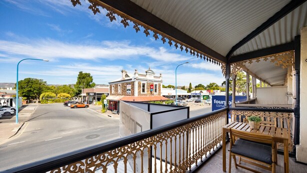 Un balcone con ringhiere in metallo decorate, una panca in legno e vista su una strada con edifici e cielo sereno.