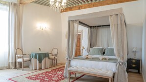 A bedroom with a four-poster bed, a red and white patterned rug, a wooden bench, a nightstand with a lamp, and a table with chairs. - La chambre des Remparts Romantique (Rochemaure)