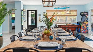 A dining area with a long wooden table set for six, surrounded by chairs. There is a pool table and a colorful arcade machine in the background. - 6BR Htd Pool, Tiki Hut, Mini golf, 8 min to Beach (Bradenton)