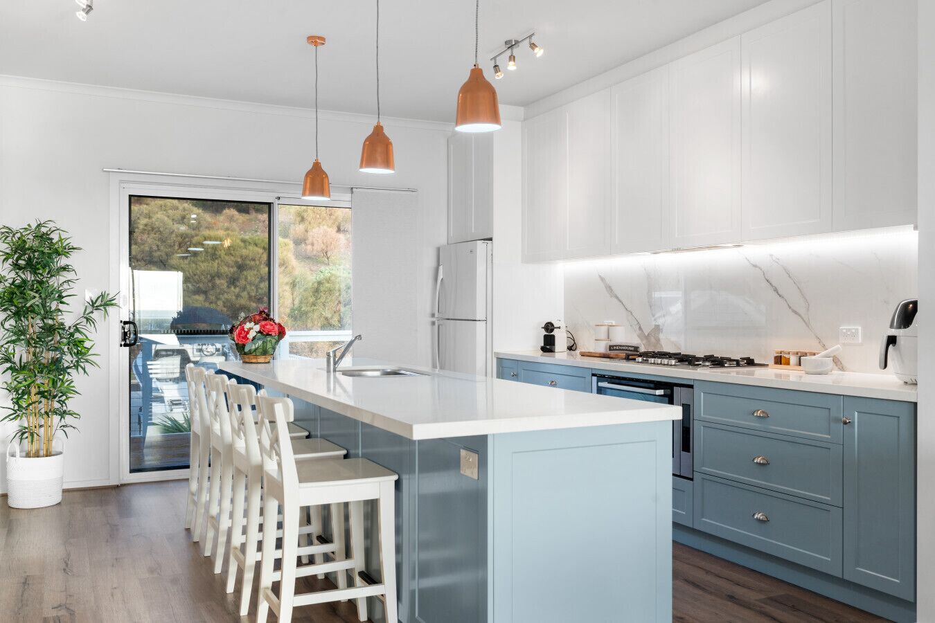A modern kitchen with a large island, pendant lights, and a view of a forest through a sliding door.