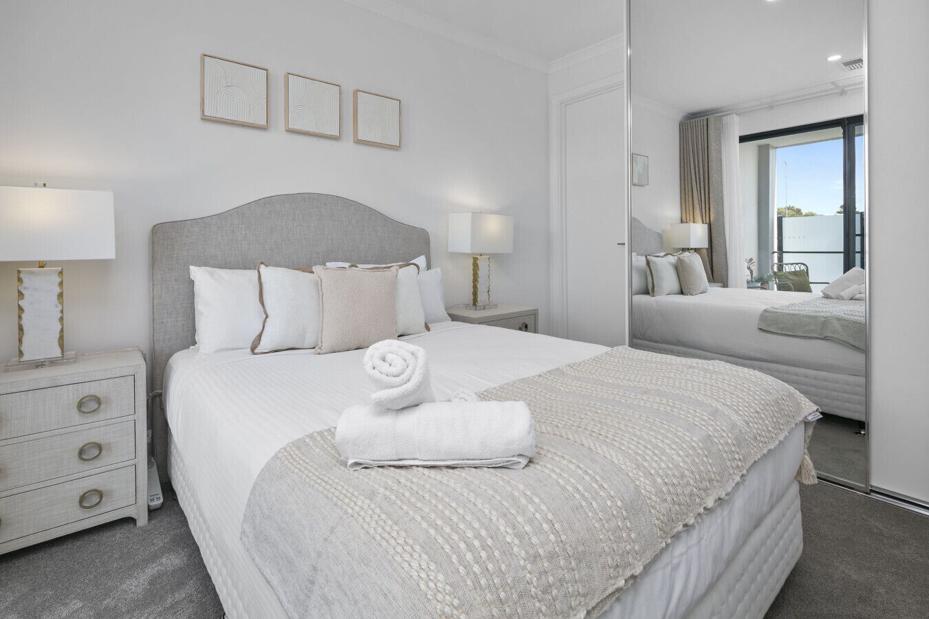 A modern hotel room with a large bed, a bedside table, a mirror, and a view of the outdoors.