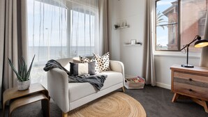 A modern living room with a sofa, a small side table, a lamp, and a view of the sea through large windows. - Horizon 235—Seaview Hideaway on the Esplanade (Glenelg North)