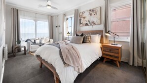 A bedroom with a large bed, bedside tables, a ceiling fan, and a window with curtains. - Horizon 235—Seaview Hideaway on the Esplanade (Glenelg North)
