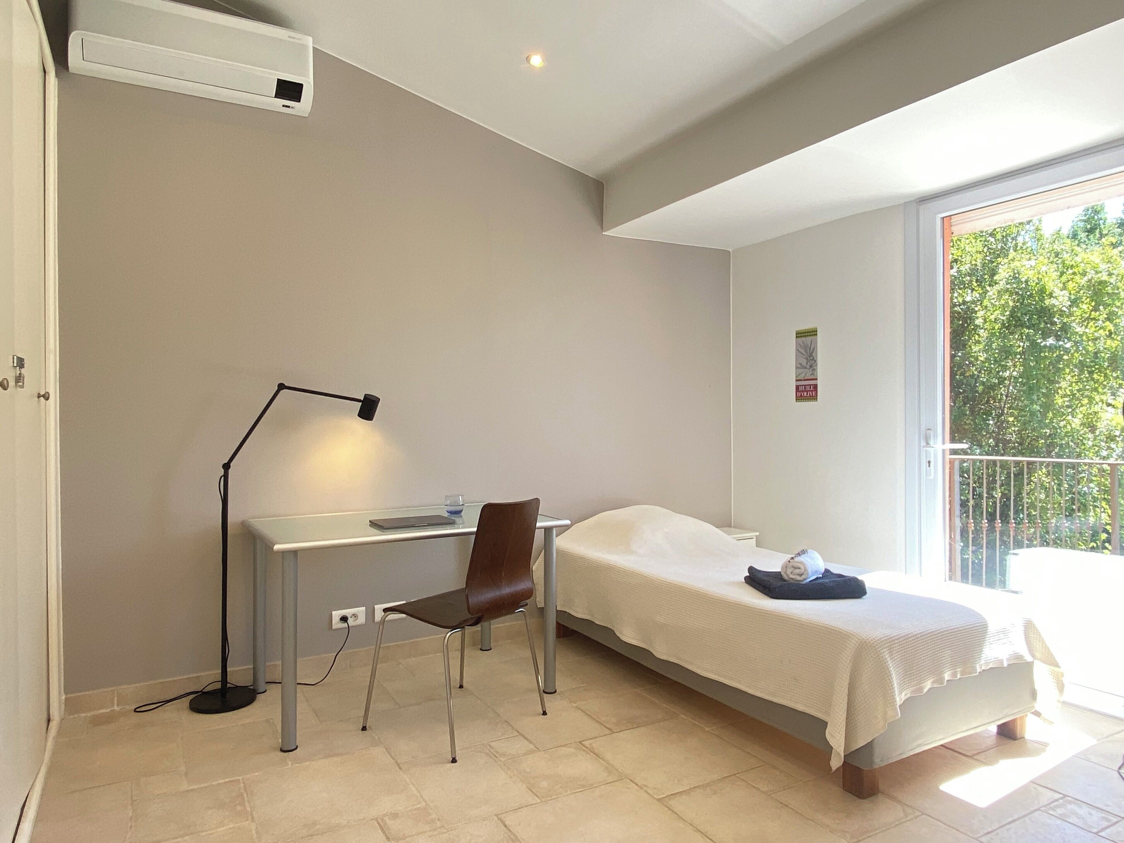 A bedroom with a bed, a desk, and a chair. There is a window with a view of trees.