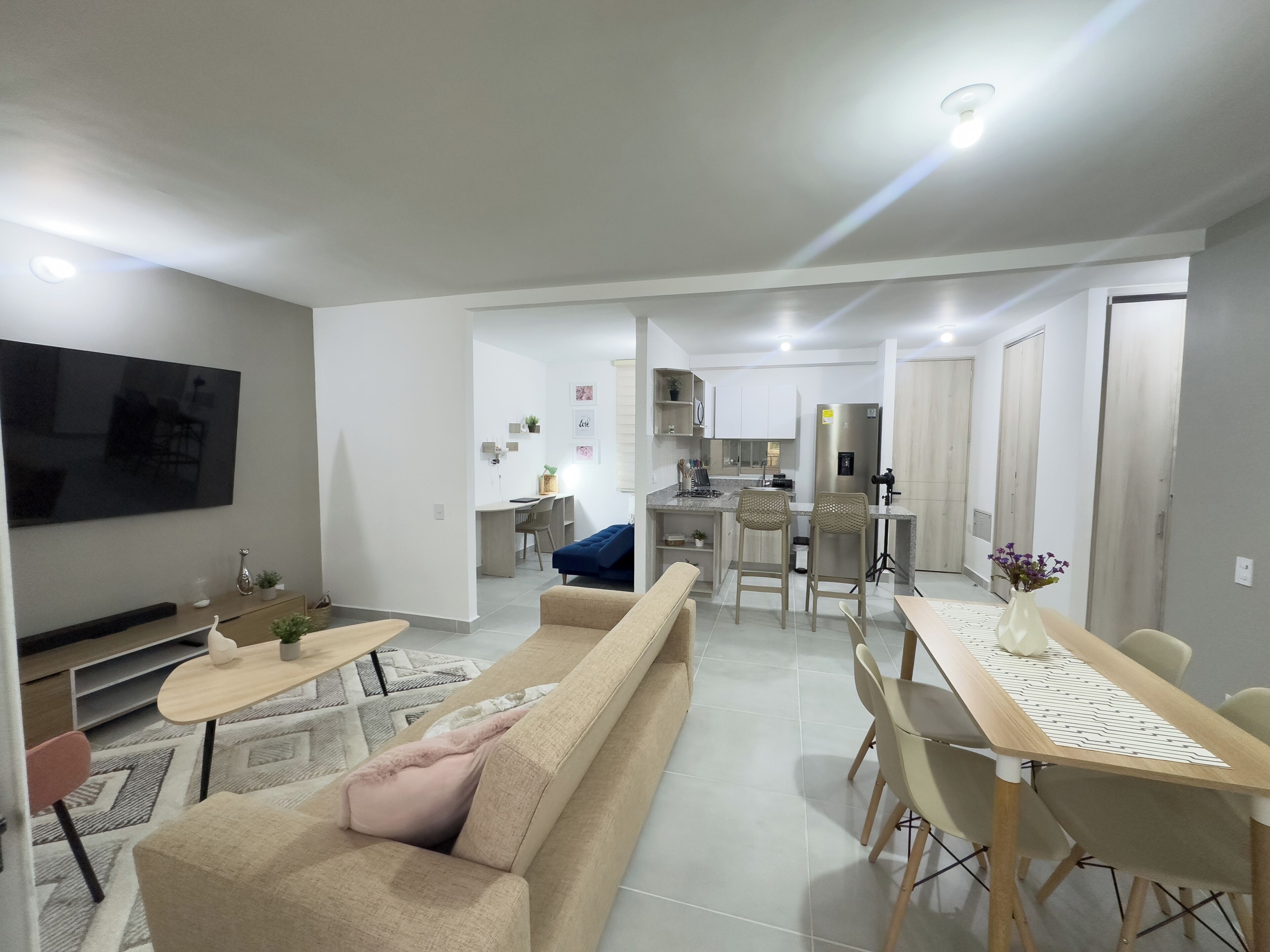 Un salon moderne avec un canapé, une table basse et un téléviseur. On aperçoit une cuisine avec un coin repas à l’arrière-plan.