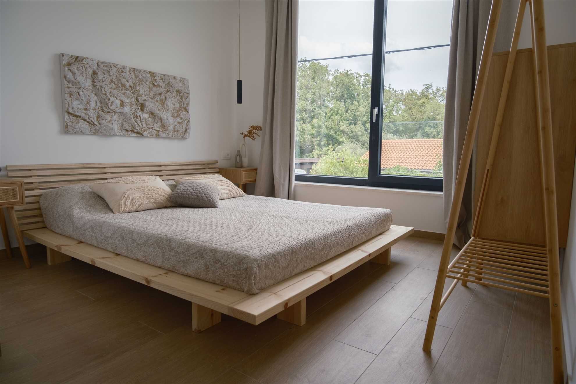 A bedroom with a wooden bed, a large window with a view of greenery, and a wooden ladder.