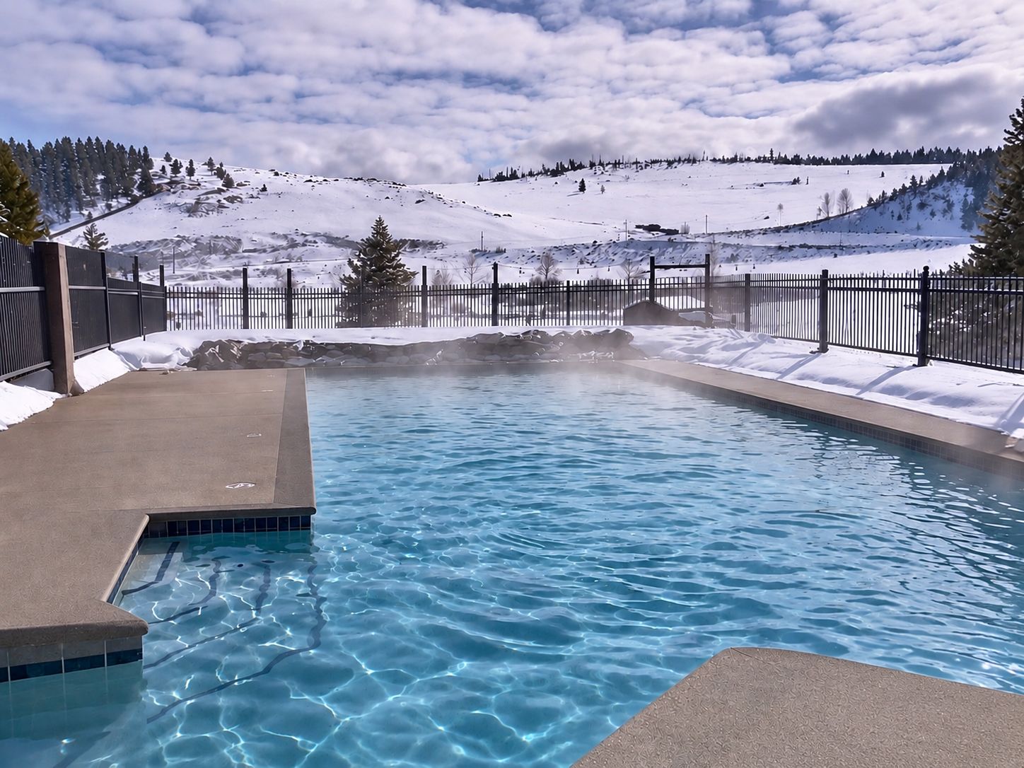 A swimming pool with clear water, surrounded by a snowy landscape and a metal fence.