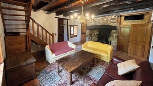 Ein rustikales Interieur mit freiliegenden Holzbalken, einem Kamin aus Stein, einem gelben Sofa und einer Holztreppe.