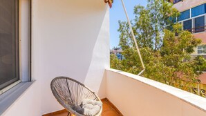 A balcony with a wicker chair, a window, and a view of buildings and trees.