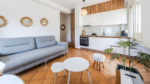 A modern living room with a sofa, two small tables, and a coffee table. There is a kitchen area with white cabinets and a wooden countertop.