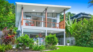 A modern two-story house with a balcony, surrounded by lush greenery and a well-maintained lawn. - Sandveil | Absolute Beachfront Access Home with Pool, Ocean Views & Family Space (Trinity Beach)