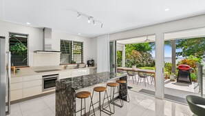 A modern kitchen with a marble island, bar stools, and a view of the outdoors. - Sandveil | Absolute Beachfront Access Home with Pool, Ocean Views & Family Space (Trinity Beach)