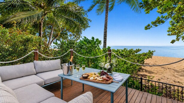 A deck with a sofa, a table with food and drinks, and a view of the beach. - Sandveil | Absolute Beachfront Access Home with Pool, Ocean Views & Family Space (Trinity Beach)