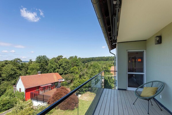 A balcony with a glass railing overlooking a red-roofed house and dense greenery.
