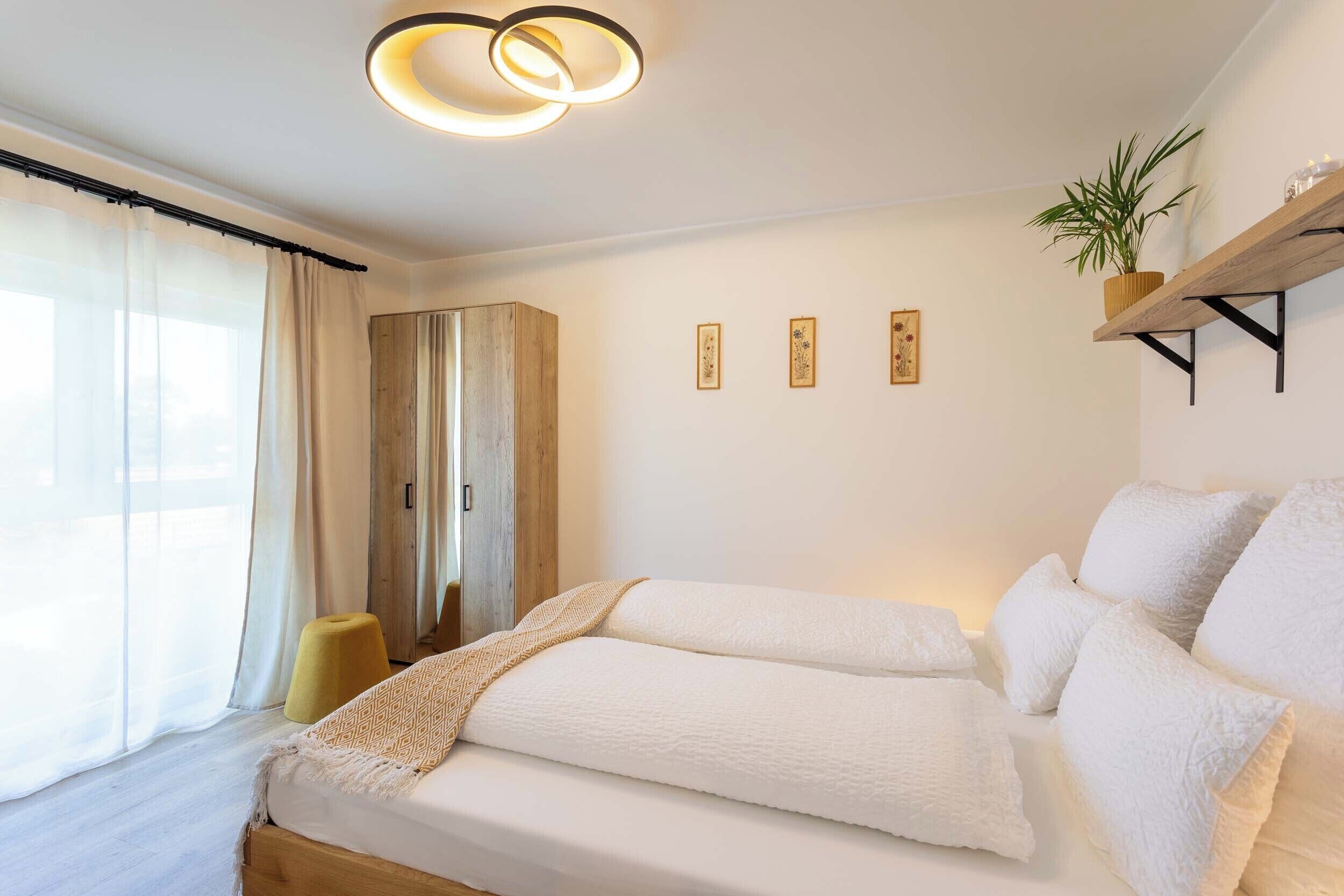 Un dormitorio con cama, armario de madera, taburete amarillo, planta en estante y cuadros en la pared.