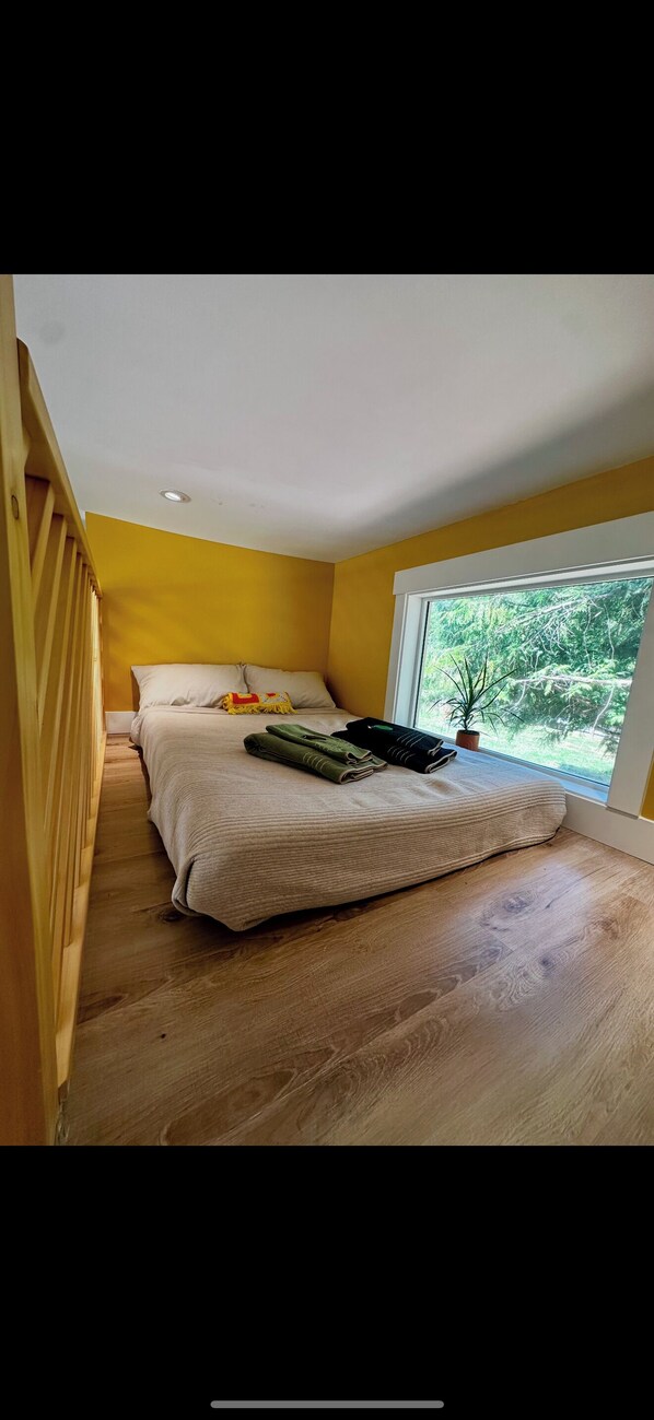 A bedroom with a large bed, a window with a view of greenery, and a wooden floor.