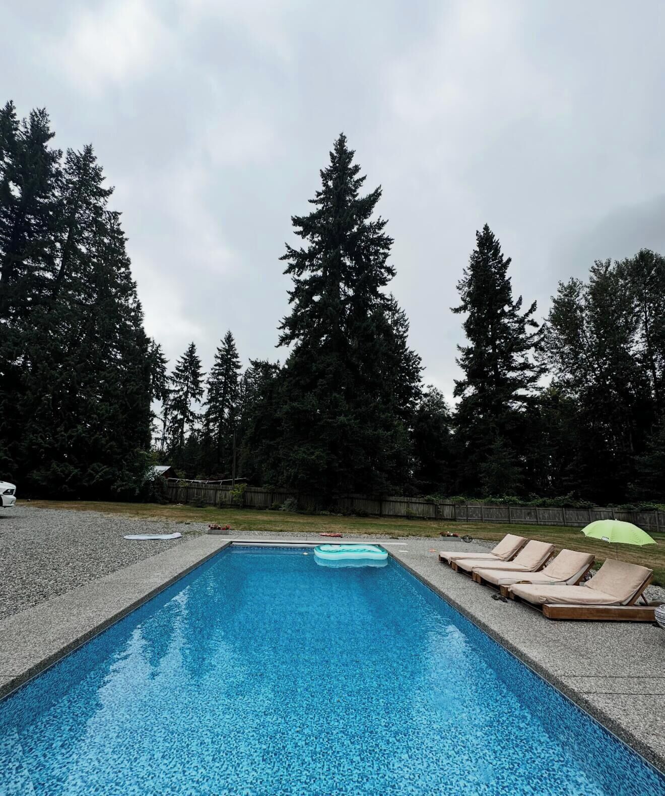 A rectangular swimming pool with clear blue water, surrounded by a pebble border. There are lounge chairs along the pool's edge, and a green umbrella is visible in the background.