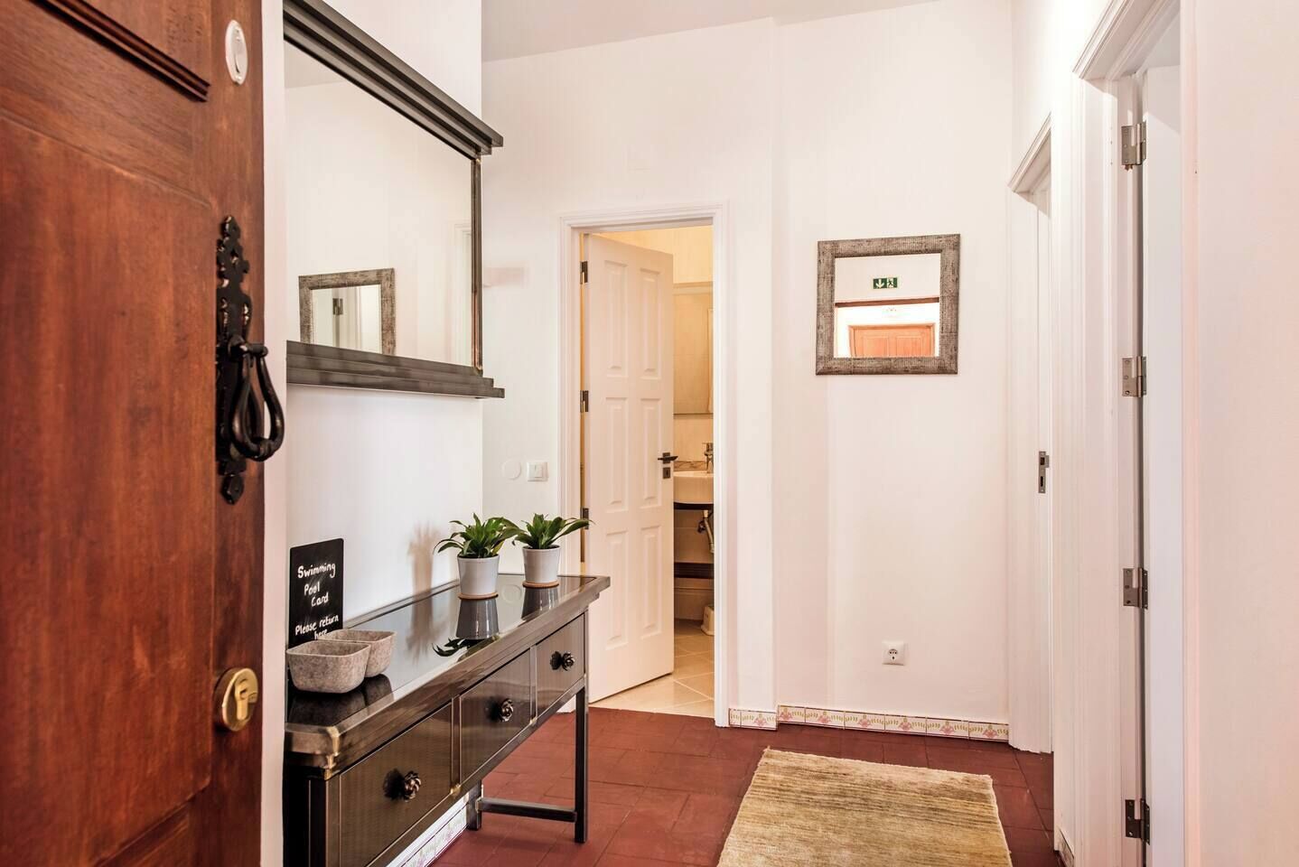 A hallway with a wooden door, a mirror, a shelf with plants, a sign, and a door with an emergency exit sign.