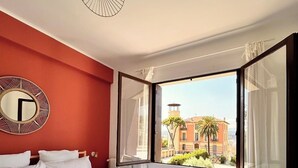 A hotel room with a large bed, a view of buildings through the window, and a round mirror on the wall. - Near Port & City - Private Parking & Breakfast (Nice)