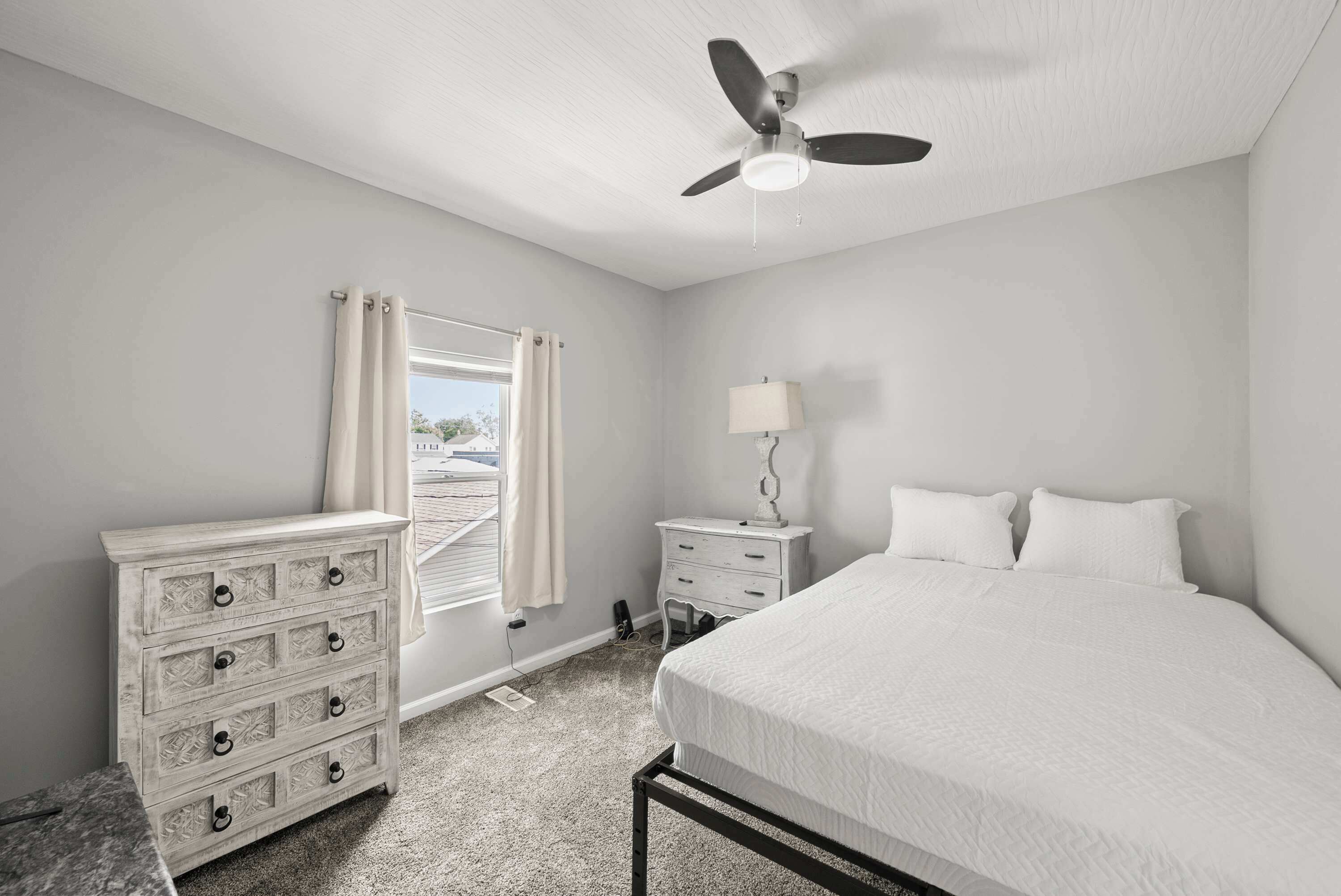 A bedroom with a bed, a dresser, a nightstand, a ceiling fan, and a window with curtains.
