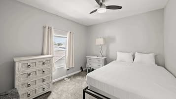 A bedroom with a bed, a dresser, a nightstand, a ceiling fan, and a window with curtains.