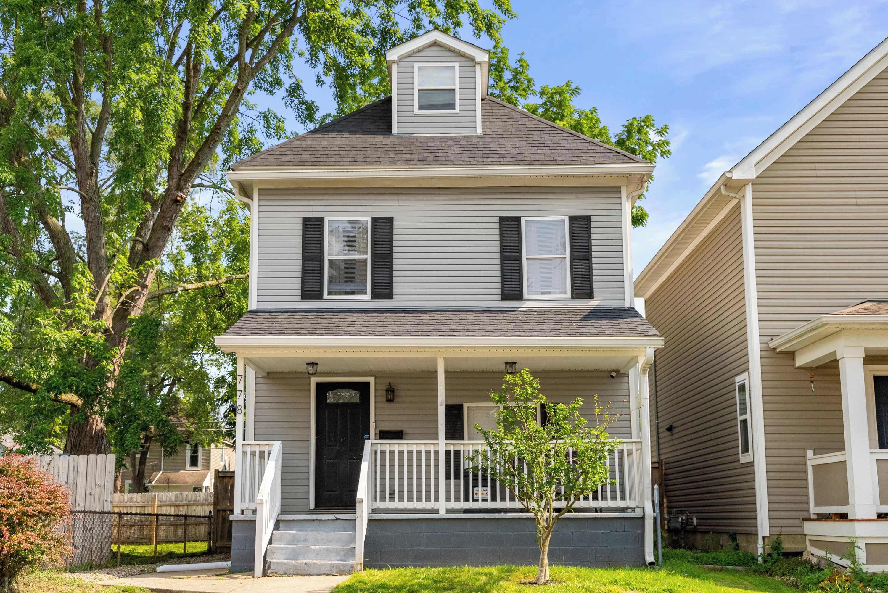A two-story house with a front porch, a small tree in front, and a fence in the background.