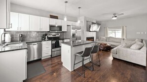 A modern kitchen with stainless steel appliances, a central island with bar stools, and a grey sofa in a living area with a fireplace. - The Southern Nook - For 8 (Columbus)