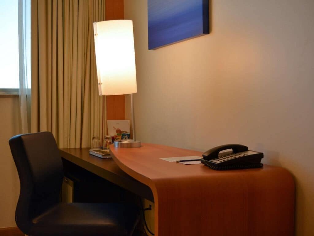 A hotel room with a desk, a lamp, a telephone, and a chair.