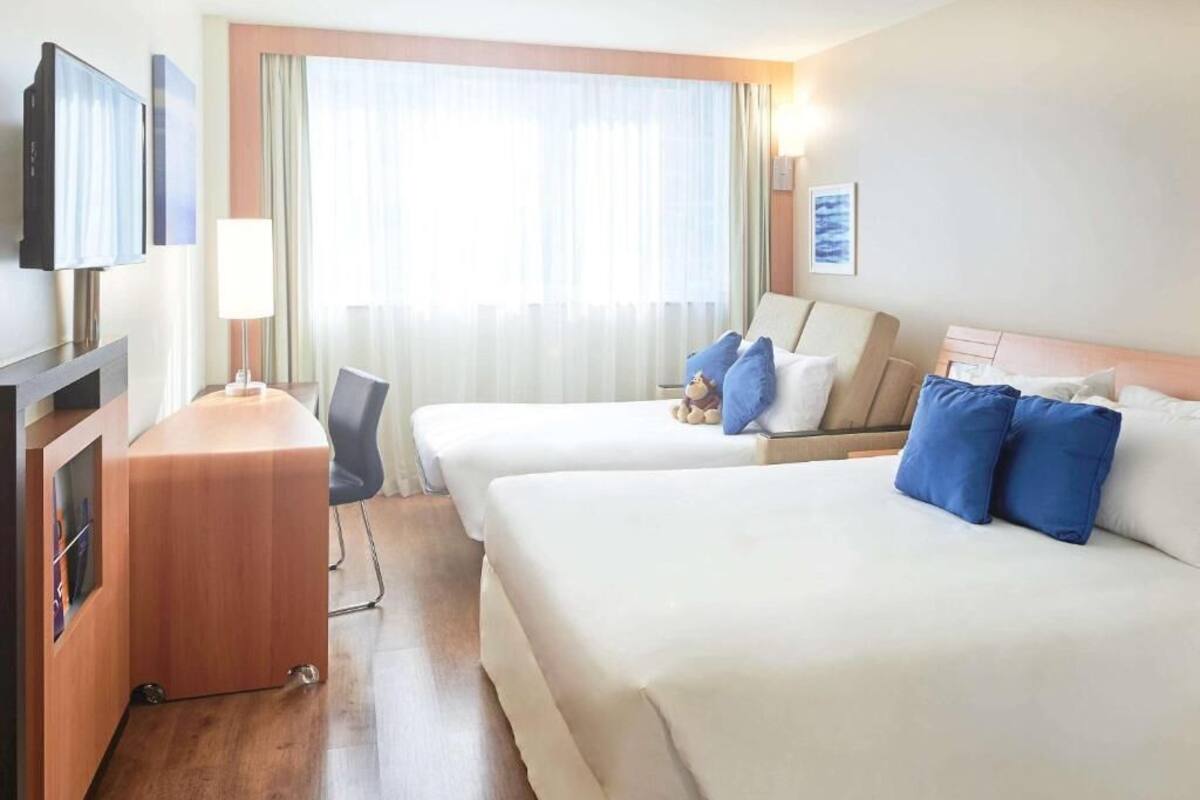 Una camera d'albergo con due letti, una scrivania, una sedia, una TV e una finestra con tende.