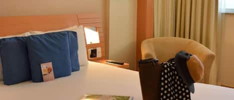 Una habitación de hotel con cama, almohadas, una silla, una lámpara y un libro.