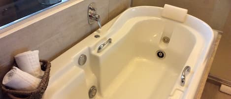 Una bañera grande y rectangular con dos llaves y desagüe, una cesta pequeña con toallas y una ventana sobre la bañera.