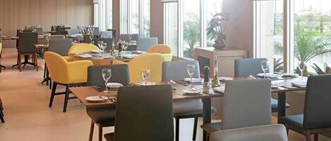 A spacious dining area with multiple tables and chairs, large windows providing natural light, and a view of greenery outside.