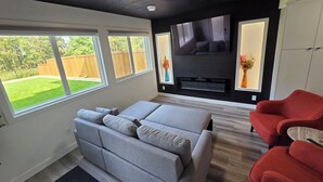 A modern living room with a grey sofa, red armchairs, a flat-screen TV, and a fireplace. - The Walkout at the Ravine (Edmonton)