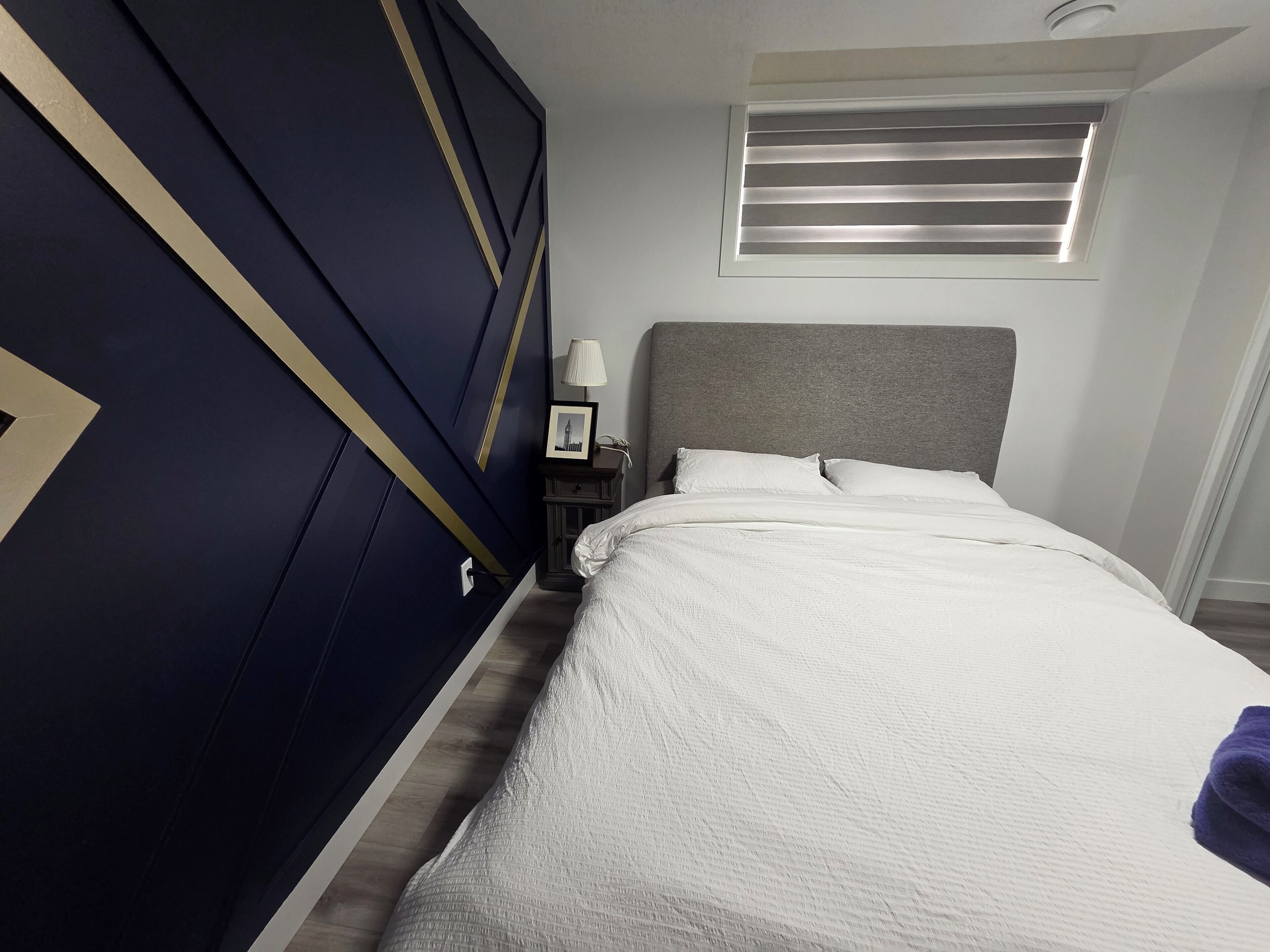 Ein Schlafzimmer mit Bett, Nachttisch, Lampe und einem Fenster mit Jalousien.