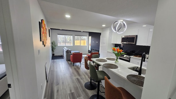 A modern kitchen with white cabinets, a stainless steel microwave, and a black stove. There is a dining area with a white table and red chairs. - The Walkout at the Ravine (Edmonton)
