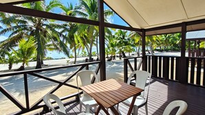 A covered outdoor seating area with a wooden table and white plastic chairs, overlooking a beach with palm trees and a clear blue sky. - Sunset Bugalows (Aitutaki)