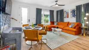 A modern living room with a sofa, chairs, a dining table, and a TV mounted on the wall.