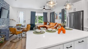 A modern kitchen with white cabinets, a large island, and stainless steel appliances. There is a dining area with a table and chairs, and a living room with a sofa and a TV.
