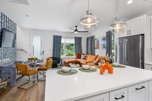 Uma cozinha moderna com armários brancos, uma ilha grande e eletrodomésticos de aço inoxidável. Há uma sala de jantar com mesa e cadeiras, e uma sala de estar com sofá e televisão.
