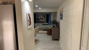 A narrow hallway with a refrigerator on the left, a bed with a blue curtain in the background, and a wooden cabinet with a plant on the right. - Spacious Studio in Heart of Harvard Square! (Cambridge)