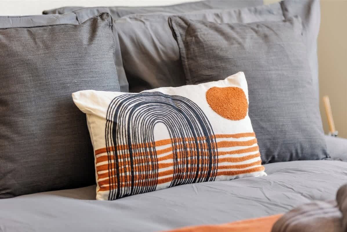 - Decorative pillow with abstract design in warm orange and black  
- Styled on gray bedding to create a modern and cozy guest room  
- Airbnb rental detail adding comfort and personality to the stay  