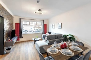 A modern living room with a dining area, a brick accent wall, a large window, and a sofa set.