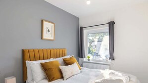 A bedroom with a bed, pillows, a nightstand, a framed picture, a window with curtains, and a radiator. - Spacious House with Private Entrance (London)