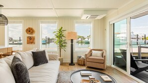 Ein modernes Wohnzimmer mit Sofa, Sessel und Couchtisch, großen Fenstern mit Blick auf eine Marina und einer Stehlampe.