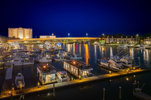 A marina at night with boats docked, a bridge in the background, and buildings on the far side.
